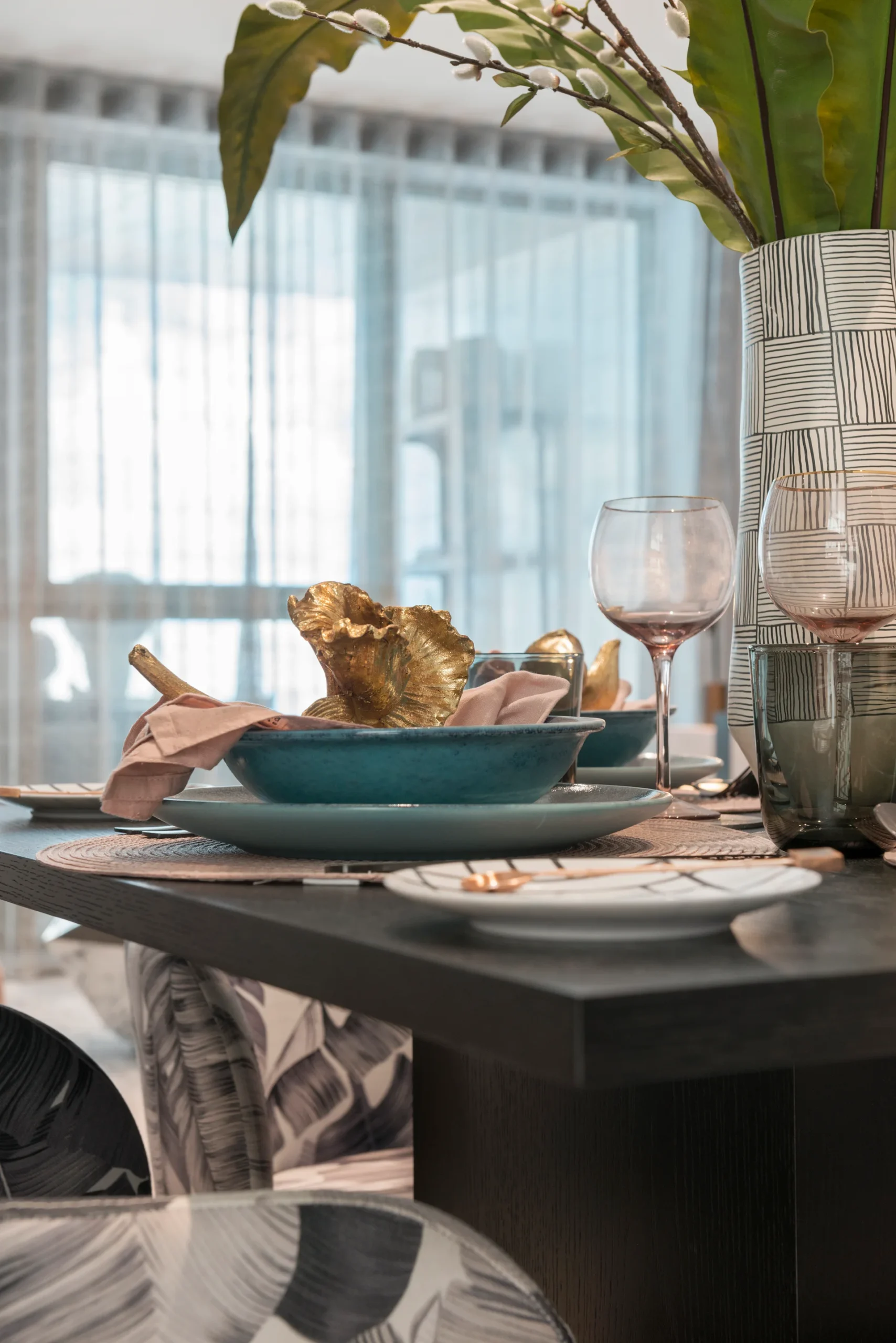 Elegant dining table set with blue dish, gold accents, wine glasses, and a decorative vase. Natural light through sheer
