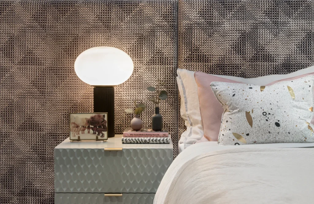 Stylish bedroom scene featuring a bedside table, lamp, and decorative pillows against patterned wallpaper.