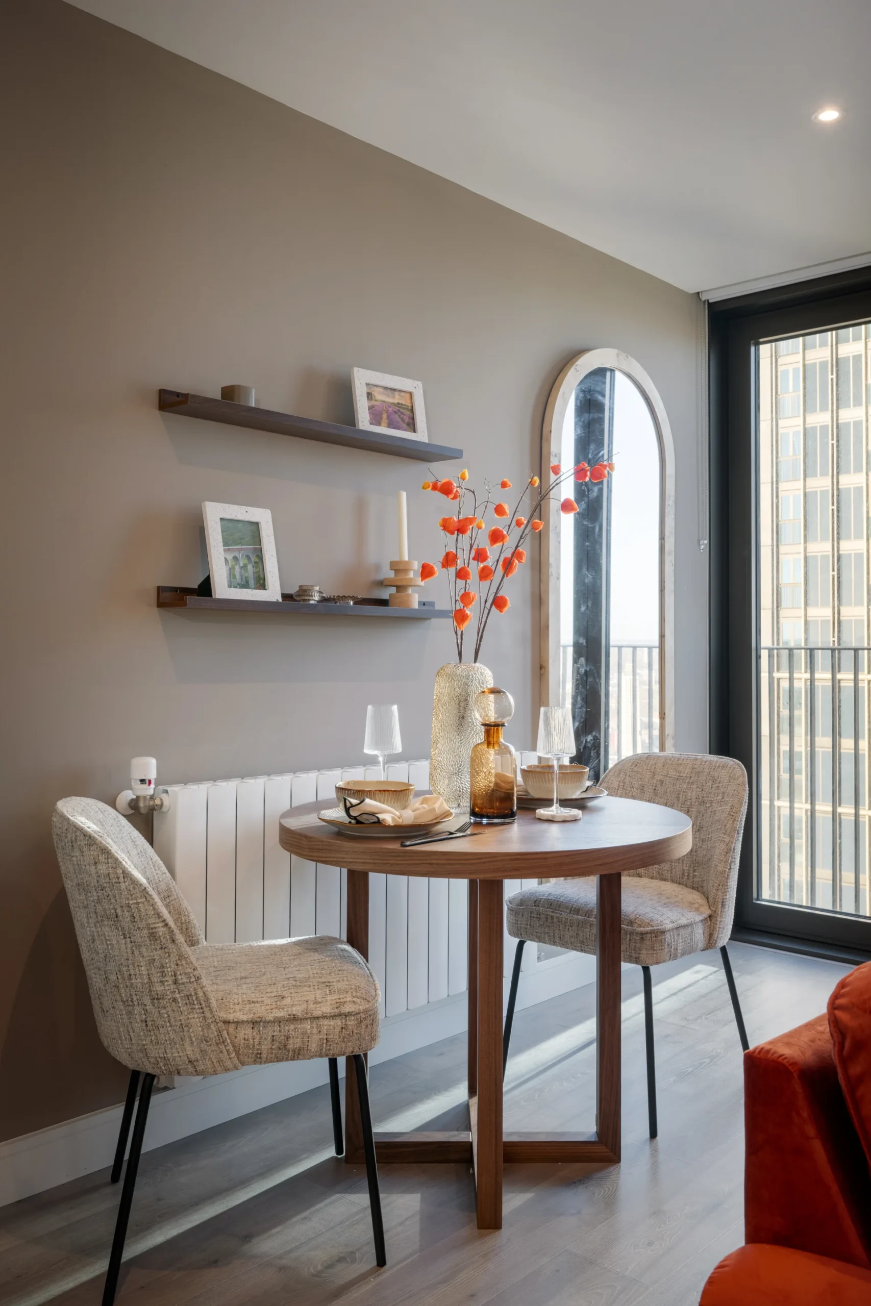 Cozy dining area with a round table, two chairs, wall shelves, and a decorative vase with orange flowers.