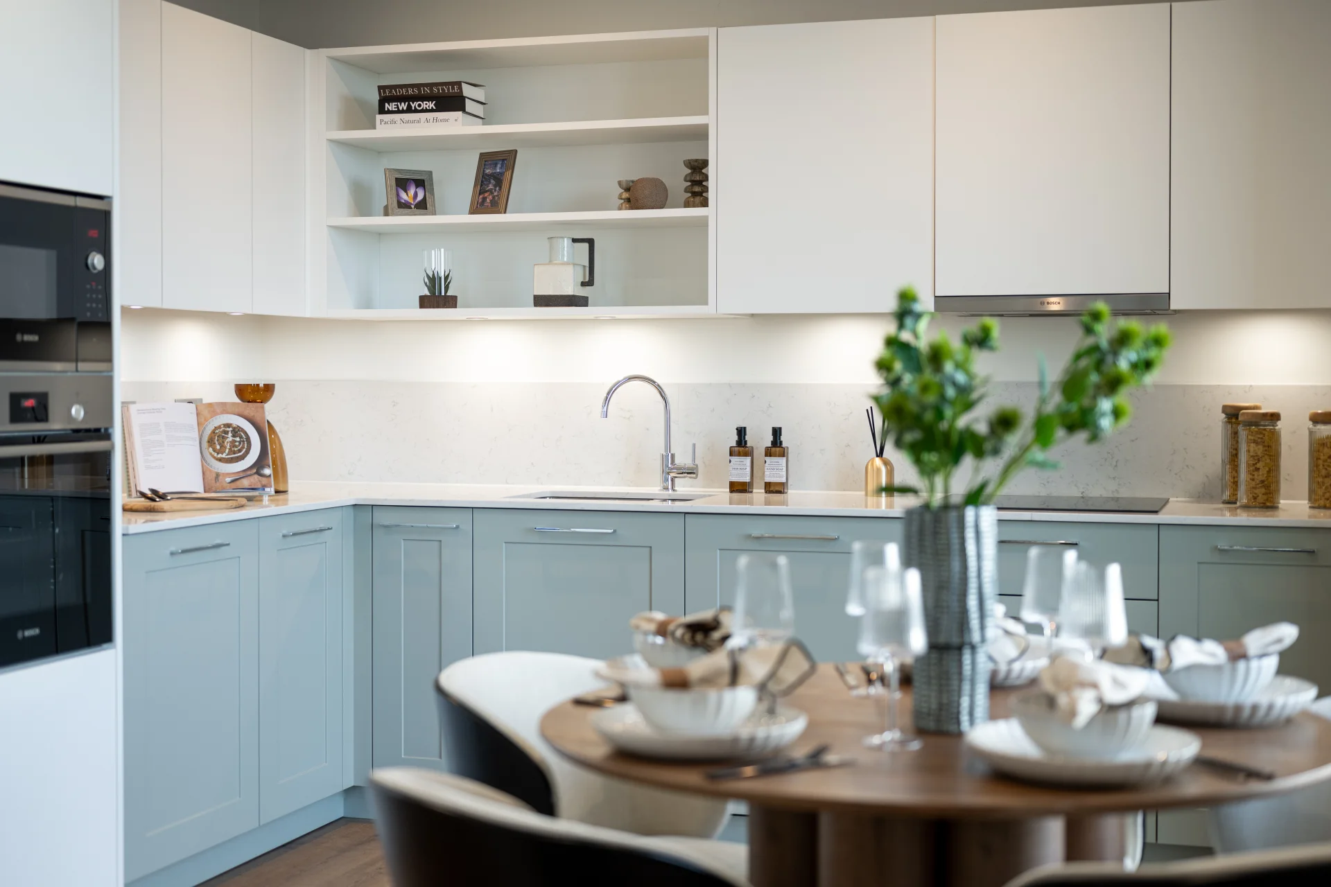 Modern kitchen with light-colored cabinetry, a wooden dining table set for a meal, and decorative plants.