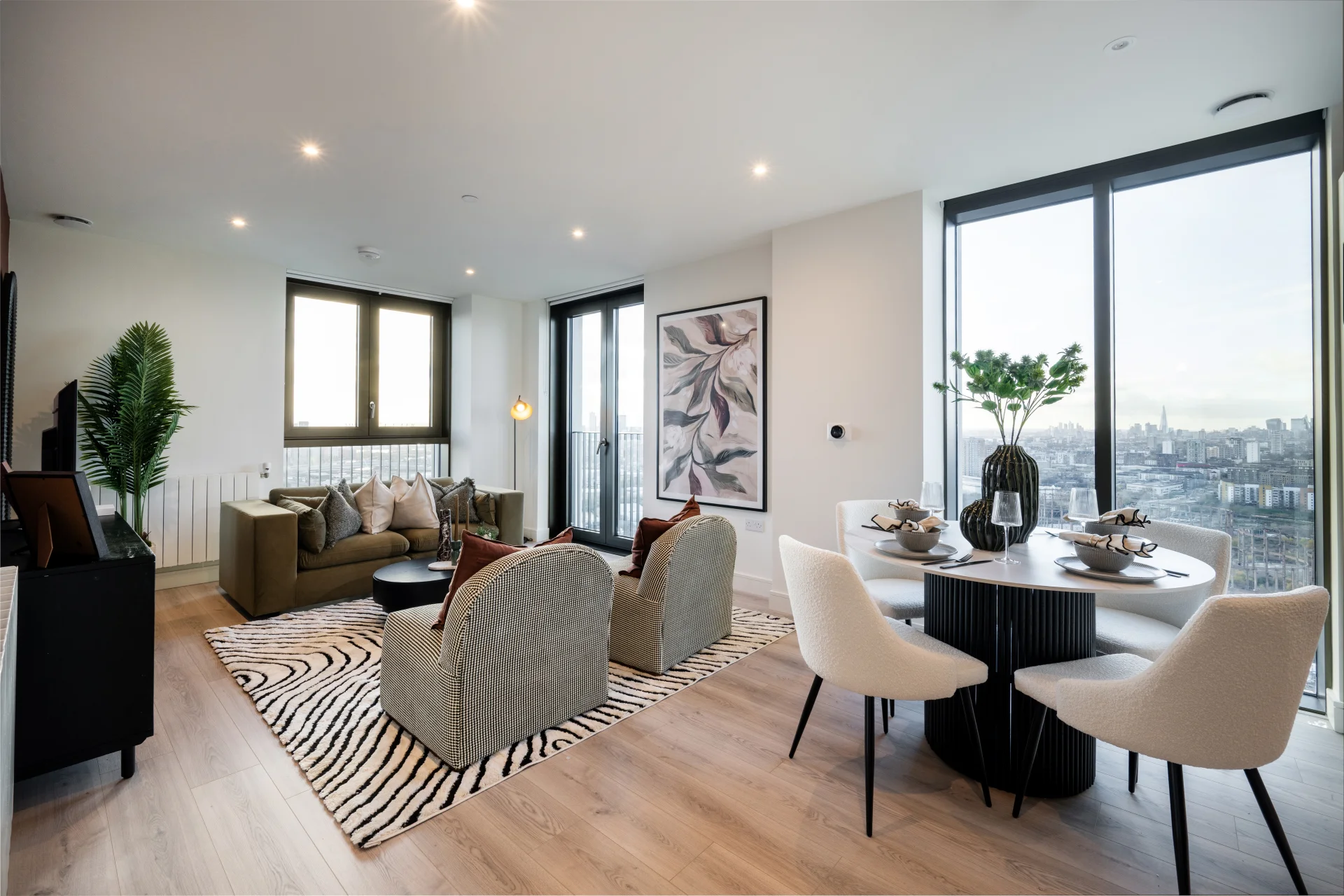 Modern living room with a dining area, plants, and large windows showcasing a city view.