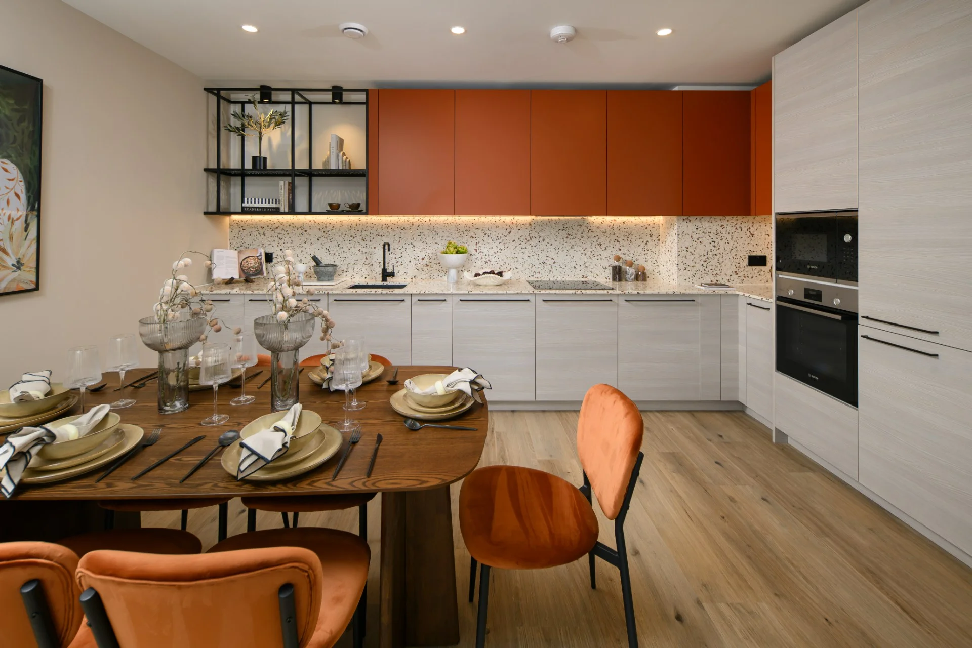 Modern dining area with a wooden table set for dinner, orange chairs, and a stylish kitchen in the background.