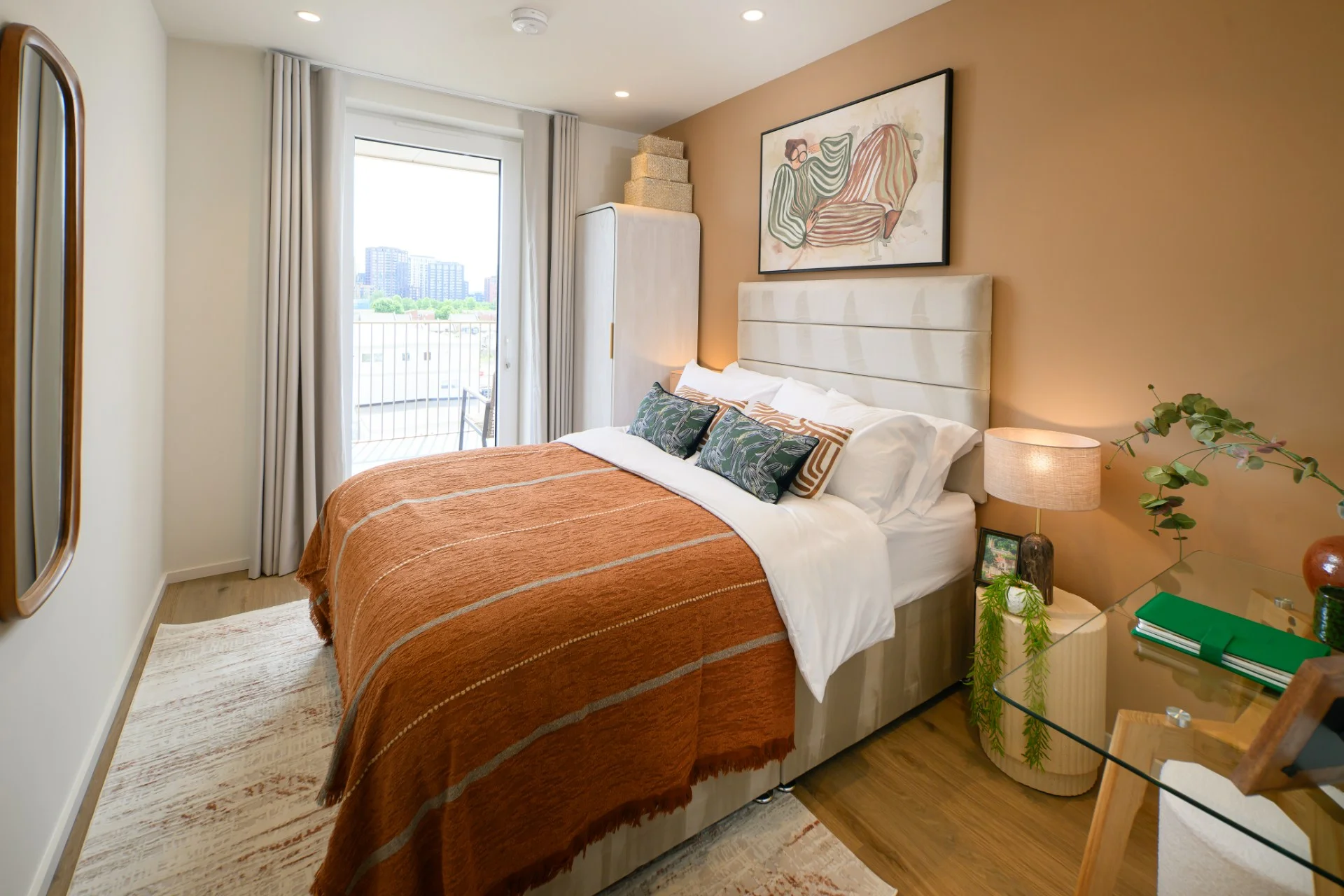 Cozy bedroom featuring a large bed with an orange blanket, decorative pillows, and a glass desk by a window.