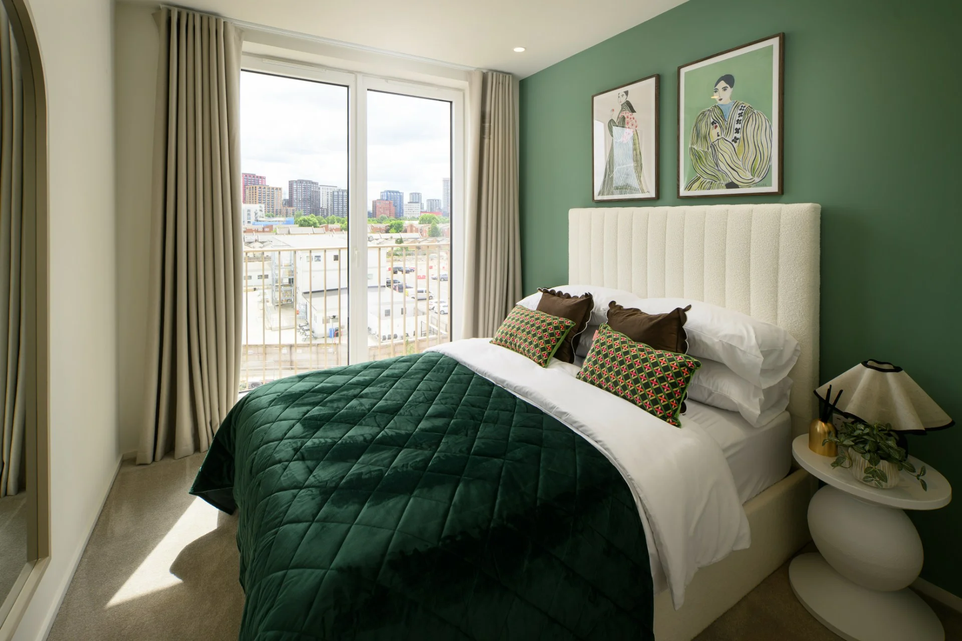 Cozy bedroom with a green accent wall, a plush bed with patterned cushions, and large windows overlooking the city.